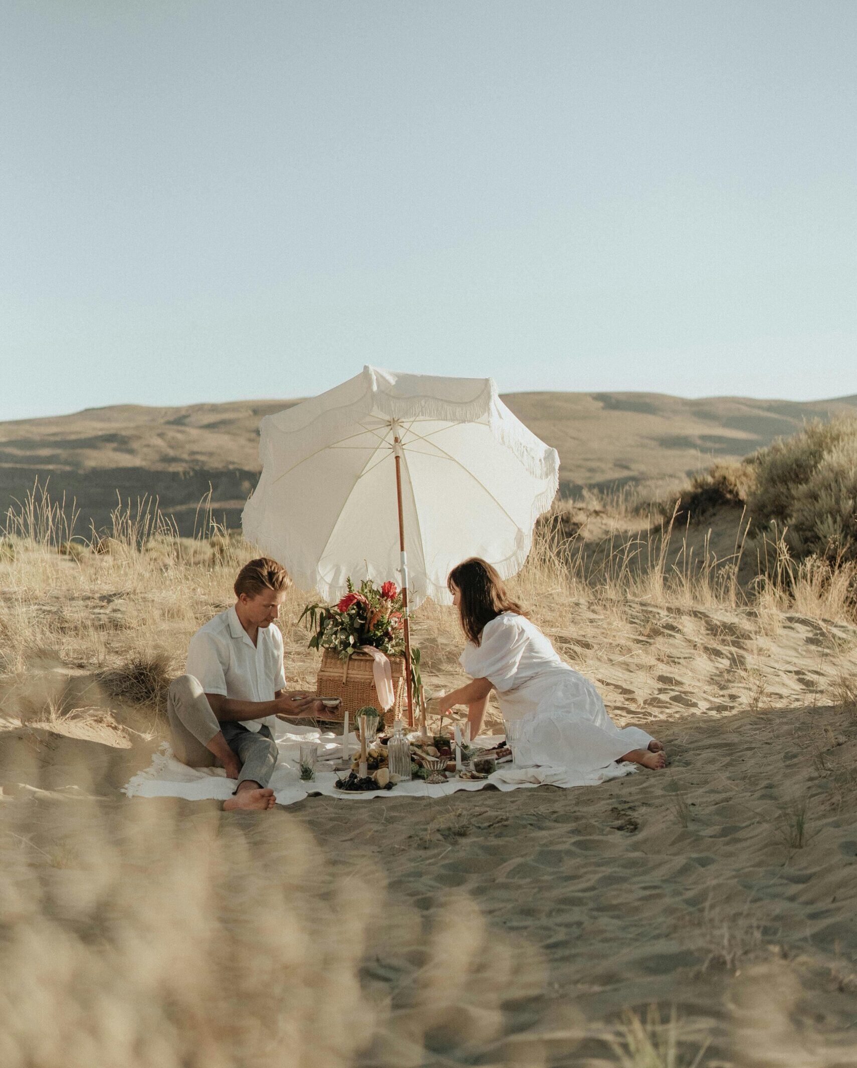 Young couple in white clothes sitting together on sandy dune and having picnic under white umbrella